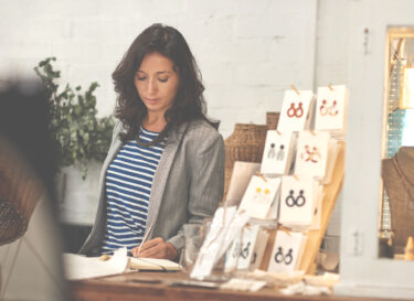 Business woman standing behind a counter in her trendy boutique writing down sales tax numbers in a notebook.