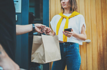 Man courier delivers bag of food to woman with a phone.