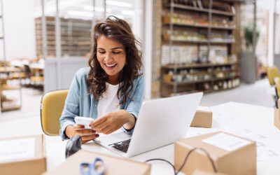 Woman managing her inventory in the back office with a computer and a cellphone surrounded by boxes.