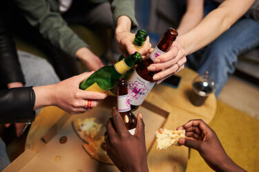 Close up shot of people hands toasting with beer bottles at home party with pizza.