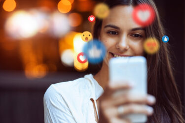 Woman holding a cell phone in front of her face. Social media emojis are swirling up from the screen.