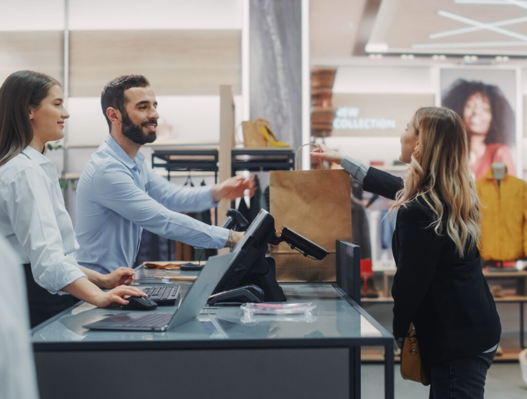 New retail employee shadowing another retail clerk and helping a woman with a purchase.