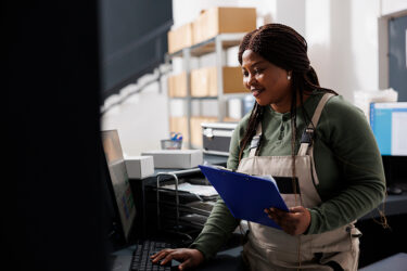 Woman managing her inventory holding a clipboard and working on a computer. She is in the back office using a ERP solution and POS software to track inventory.