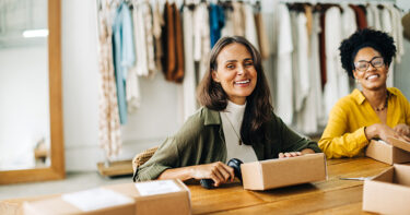 Two women in the back office of a retail store performing a year-end inventory count. There is a rack of clothing in the background.